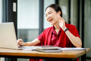 Portrait of Cheerful attractive Asian young business woman working on laptop in cafe,doing finances,accounting analysis,report,data and pointing graph and Wear stylish red suit jacket.