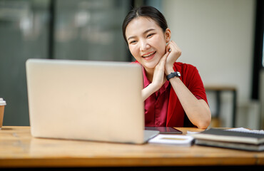 Portrait of Cheerful attractive Asian young business woman working on laptop in cafe,doing finances,accounting analysis,report,data and pointing graph and Wear stylish red suit jacket.