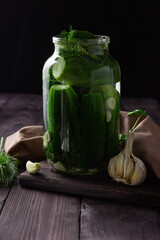 Lightly salted cucumbers in a glass jar with garlic on a dark wooden table.