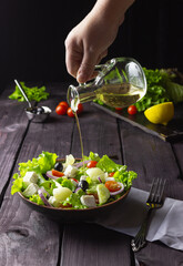 Greek salad in a plate on a cutting board on a dark wooden background. Healthy vegetarian food. A hand pours olive oil from a glass decanter.