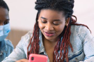 An African-American woman sits with a pink phone in her hands and another woman peers over her...