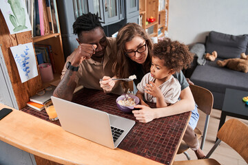 Woman feeding with breakfast her lovely multiracial child while sitting in front of laptop