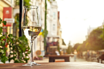 Glass of white wine close up. Alcoholic drink on a wooden table on a background of the city. Transparent wine on the summer terrace of the restaurant. Blurred people in the background.