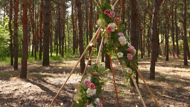 Bohemian tipi arch made of wooden rods decorated with pink roses, candles on carpet, pampass grass, wrapped in fairy lights on outdoor wedding ceremony venue in pine forest. Floristic compositions.