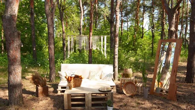 Lounge photo zone with diy sofa and table of cargo pallets in boho style on outdoor wedding ceremony venue in pine forest. Bohemian decor.