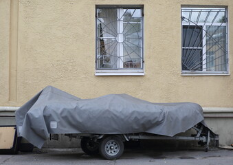 Covered with a gray cover, a long two-wheeled trailer is parked at the yellow wall, ulitsa Sedova, St. Petersburg, Russia, August 2021