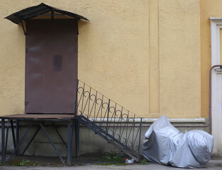 The motorcycle is covered with a cover and parked at the entrance with a metal staircase and a porch, ulitsa Sedova, St. Petersburg, Russia, August 2021