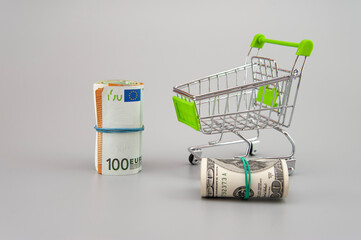 euro and dolar banknotes in rolls with supermarket trolley on gray background
