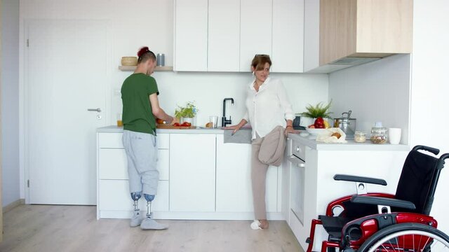 Rear view of disabled mature woman with wheelchair preparing food with son in kitchen indoors at home.