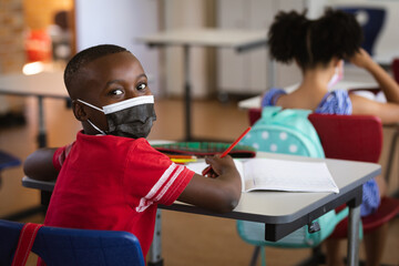 Portrait of african american boy wearing face mask sitting on his desk in class at elementary school - Powered by Adobe