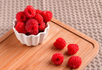 Ripe raspberries in a white cup on a wooden board. Textile background.