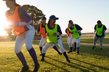 Diverse group of female baseball players wearing coloured bibs training on field at sunrise