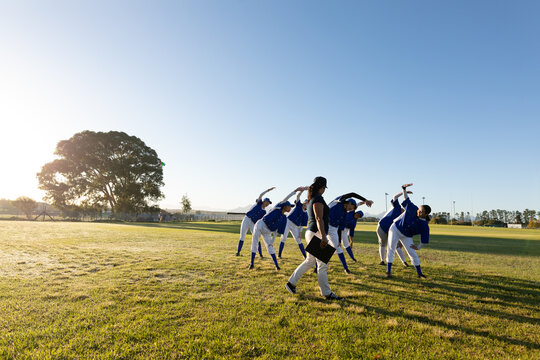 Diverse group of female baseball players with coach warming up in field, stretching from the waist