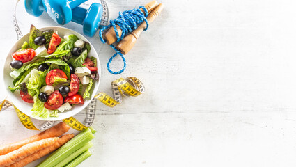 Fototapeta na wymiar Healthy fresh salad with tomatoes surrounded with exercise equipment, carrtot celery and measuring tape - top of view