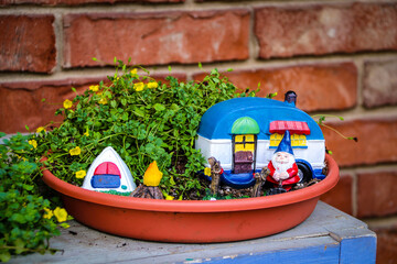 Camping gnome fairy garden in planter with minature camper and tent against blurred brick wall.