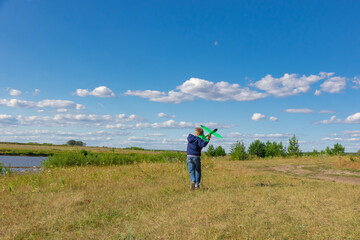 Naklejka na ściany i meble A six-year-old preschooler boy in a blue jacket launches a toy plane in a field against a blue sky with clouds on a summer day. The bright sun is shining. Scenery