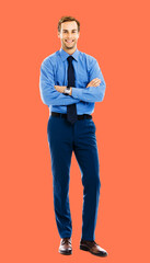 Full length body portrait of happy smiling businessman in blue cloth, standing in crossed arm pose, isolated over bright vivid orange color background. Confident business man at studio image.