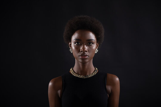 Portrait of young serious stylish black woman. Beautiful curly dark haired african american girl wear black dress with gold jewelry and looking at camera. Studio shoot isolated on dark background.