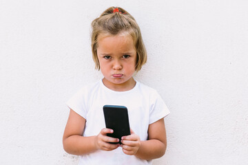 Fototapeta premium Little blond hair girl wearing white t-shirt, holding mobile phone, looking angry