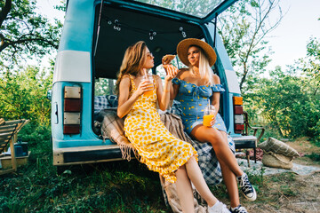 Two attractive cheerful women drinking lemonade near van and eating pizza, enjoying summer vibes in...