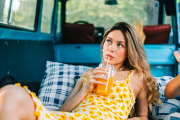 Portrait of young pretty woman sitting on blanket in a van and drinking lemonade outdoors in nature. Enjoying summer, travel concept - obrazy, fototapety, plakaty