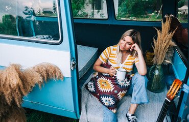 Portrait of happy young woman sitting in a van and drinking tea outdoors in nature, during sunset. Enjoying summer, travel concept