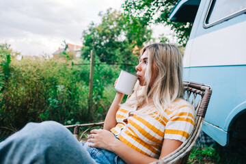 Young woman sitting on wooden chair near van and drinking tea outdoors in nature, during sunset. Enjoying summer, travel concept