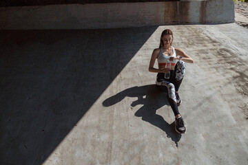 Woman in sports clothes and sneakers opens bottle of water sitting on concrete ground