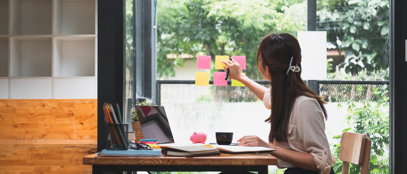 Business attractive young asian woman write notes on sticky note paper to arrange meeting with coworker sit in background take pictures through the glass, start up business and financial concept