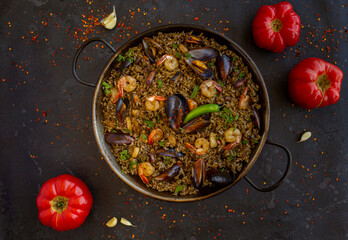 a traditional spanish black paella with shrimps, mussels and squid ink in a pan with tomatoes on a black background. view from above, flat lay