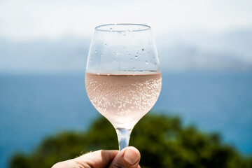 Glass of wine in hand. A glass of rose wine against the background of the Mediterranean sea and blue sky on a sunny summer day. Summer, travel, lifestyle, relaxation, and enjoyment concept.