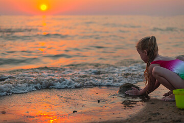 Fototapeta na wymiar little blonde girl builds a sand castle on the seashore in the evening at sunset in the golden hour in summer