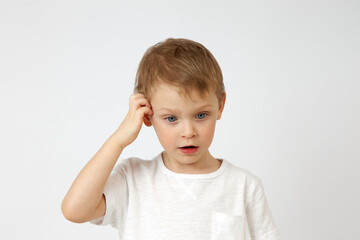 Little boy with blue eyes is deep in thought, looking down and scratching his head against a white...