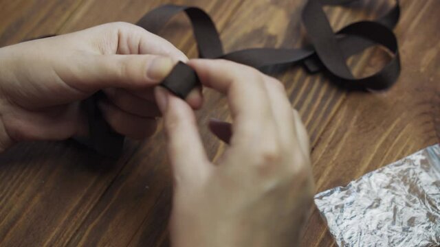 Women's hands twist brown ribbon into roll. Small package on wooden table. Workshop of craftsman or seamstress. Handmade concept.