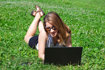 A young girl or woman lies on a meadow in the park and works on a laptop. Freelance, education or working online concept.