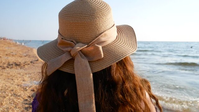 A girl in a straw hat with brown long hair looks at the sea. Rest, relaxation, travel