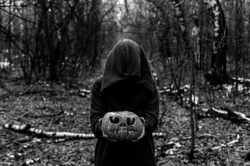 A witch holds a creepy pumpkin in a dense autumn forest. Jack o lantern for halloween. Monochrome
