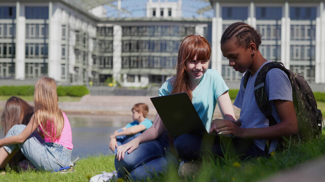 Diverse schoolchildren enjoying break on green lawn using laptop