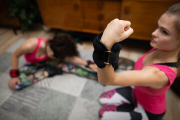 Two girls are working out, one is exercising her arm biceps muscles and the other is doing sit-ups....