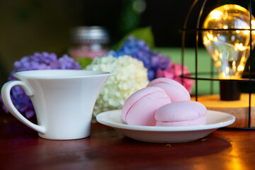 Pink marshmallow on a white saucer with multicolored hydrangea flowers and a cup against the background of evening lights. Summer evening, holiday mood concept.
