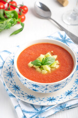 Refreshing cold gazpacho soup made of blended ripe tomatoes decorated with chopped cucumber and basil leaf served in bowl on textile napkin on white wooden background with glass of wine and spoon