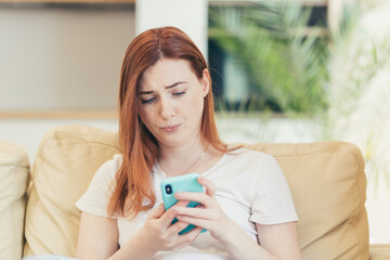 Young woman at home, frustrated and depressed reading bad news from the phone sitting on the couch