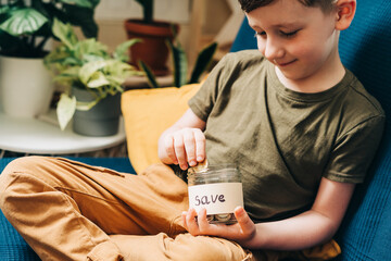 Close up of Little child kid boy hands grabbing and putting stack coins in to glass jar with save label. Donation, saving money, charity, family finance plan concept.