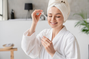 Young white woman in bath spa towel applying rejuvenation moisturizing serum on her face with her...