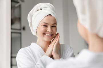 Happy white woman with toothy smile clean young clear skin in spa bath towel looking at the mirror. Beauty treatment, body and hair care. Girl after taking bath shower at home