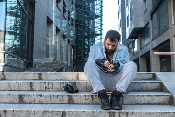 Fototapeta na wymiar Young hungry dirty depressed homeless man sitting on the stairs of the building on sidewalk and try to eat food scraps from paper bag that he find in the garbage can, social documentary concept