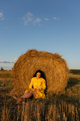Girl in yellow dress sitting near straw roll on a field.