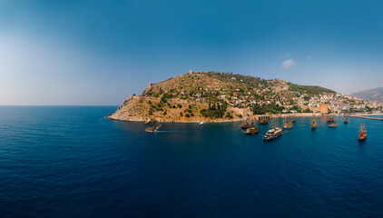 Fototapeta na wymiar Aerial wide panoramic view of Alanya in southern coast of Turkey, Summer morning day. Travel and vacation. Kalesi Castle. Ships and boats in the mediterranean. Kızılkule bay. lighthouse and pier
