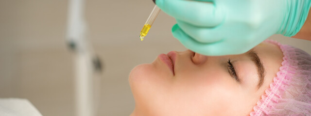 Close up of beautician holding pipette and applying oil serum drop on the face of a young woman in spa salon