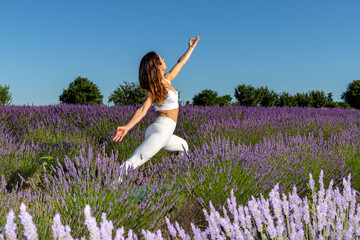 Warrior yoga pose in a blooming lavender field. A woman with long brown hair stretches the front of her body to strengthen her legs, torso and back. Sense of energy and stability.